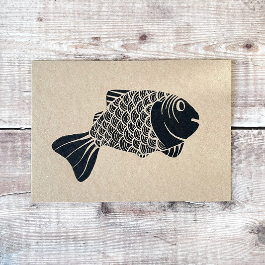 Black lino print of a fish illustration on a kraft card laid wooden surface