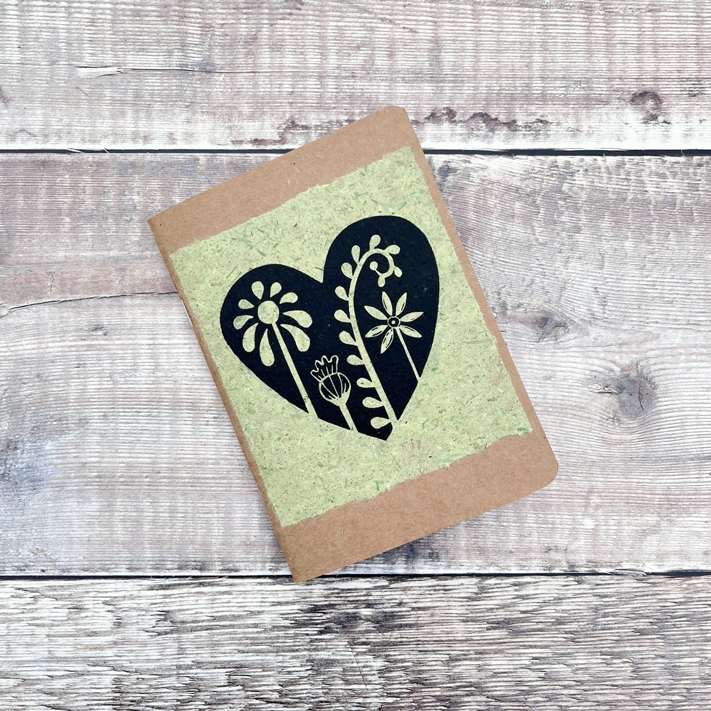 A6 kraft notebook with a black lino printed Floral Heart design on pale green handmade paper. Laid on a wooden background.