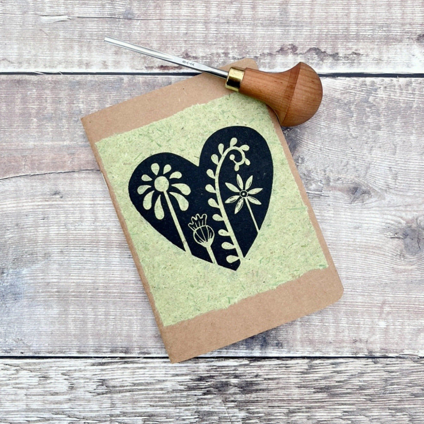 A6 kraft notebook with a black lino printed Floral Heart design on pale green handmade paper. Photo includes the lino cutting tool used, laid on a wooden background.