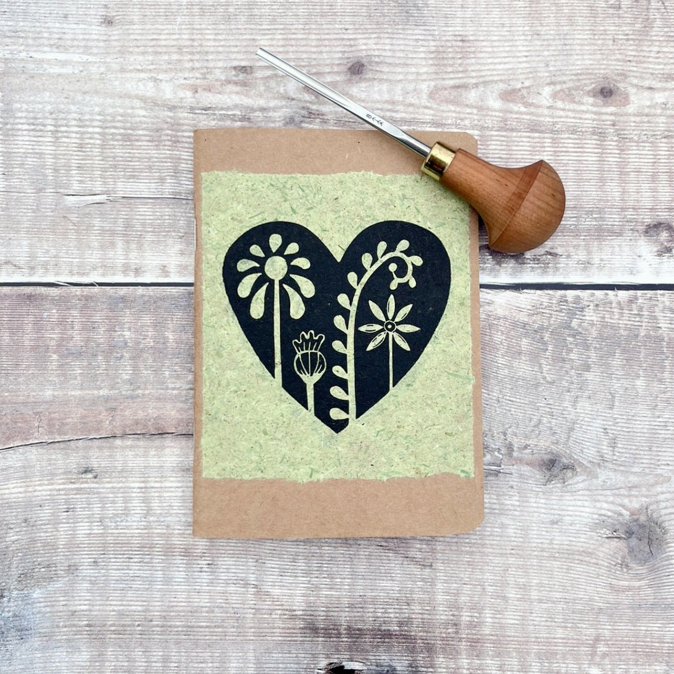 A6 kraft notebook with a black lino printed Floral Heart design on pale green handmade paper. Photo includes the lino cutting tool used, laid on a wooden background.