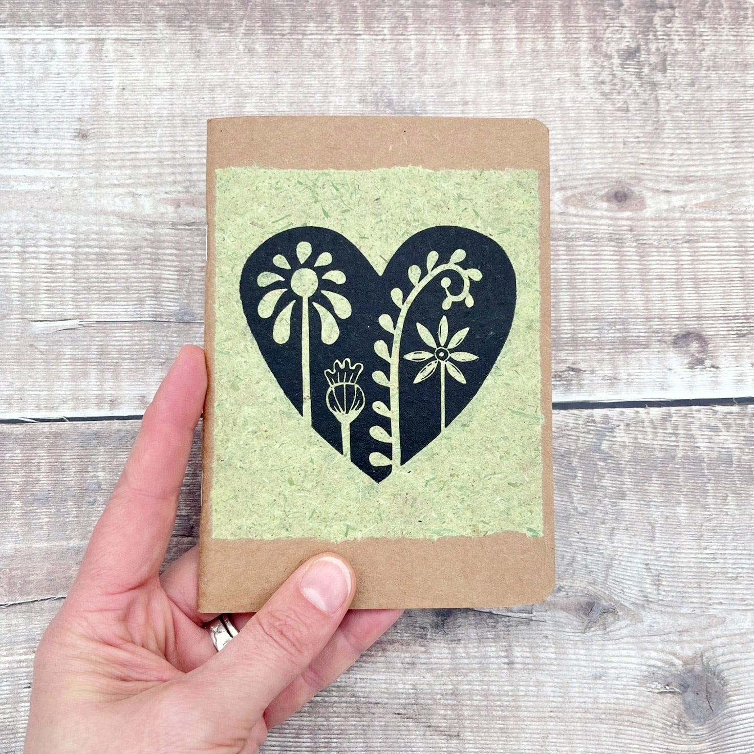 A6 kraft notebook with a black lino printed Floral Heart design on pale green handmade paper. Held in a hand against a wooden background.