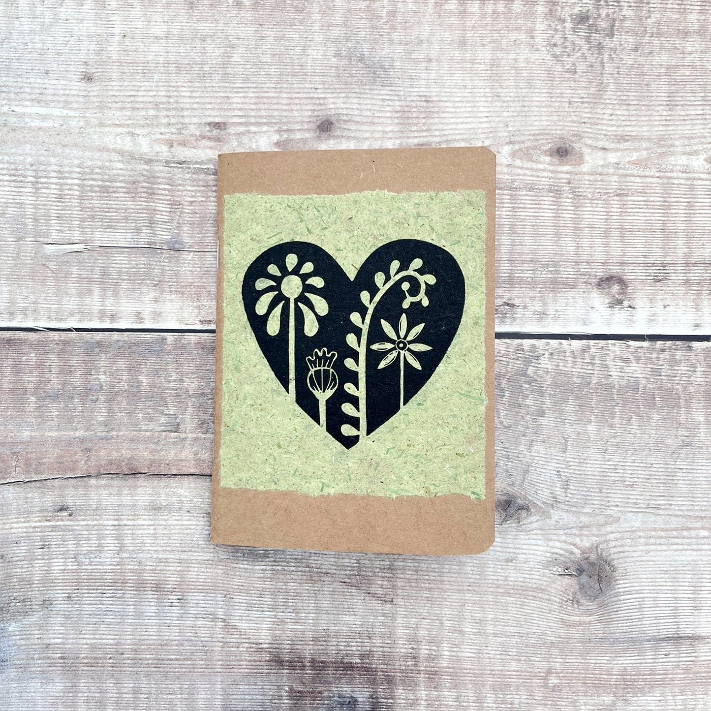 A6 kraft notebook with a black lino printed Floral Heart design on pale green handmade paper. Laid on a wooden background.