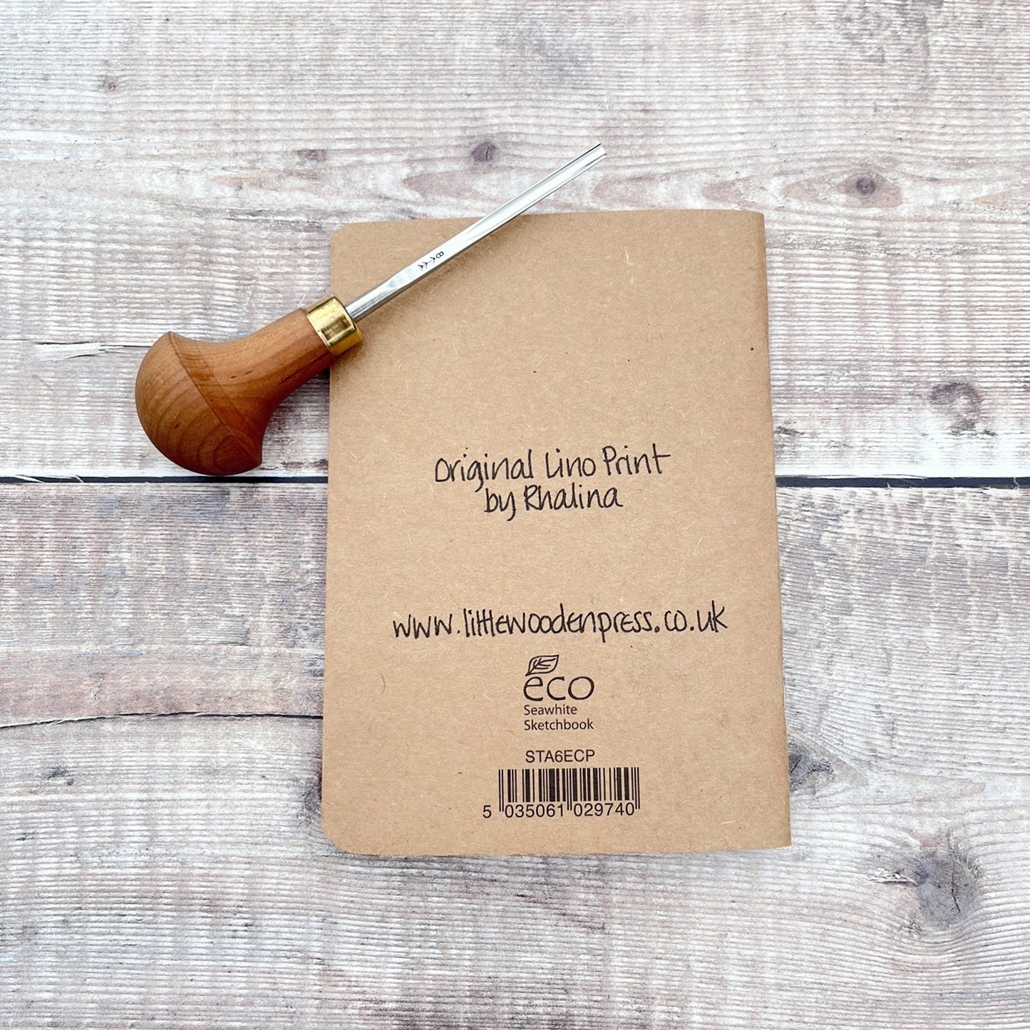 The back of the A6 kraft notebook showing the text 'Original lino print by Rhalina' www.littlewoodenpress.co.uk
Eco Seawhite Sketchbook. Photo includes lino cutting tool used. Laid against a wooden background.