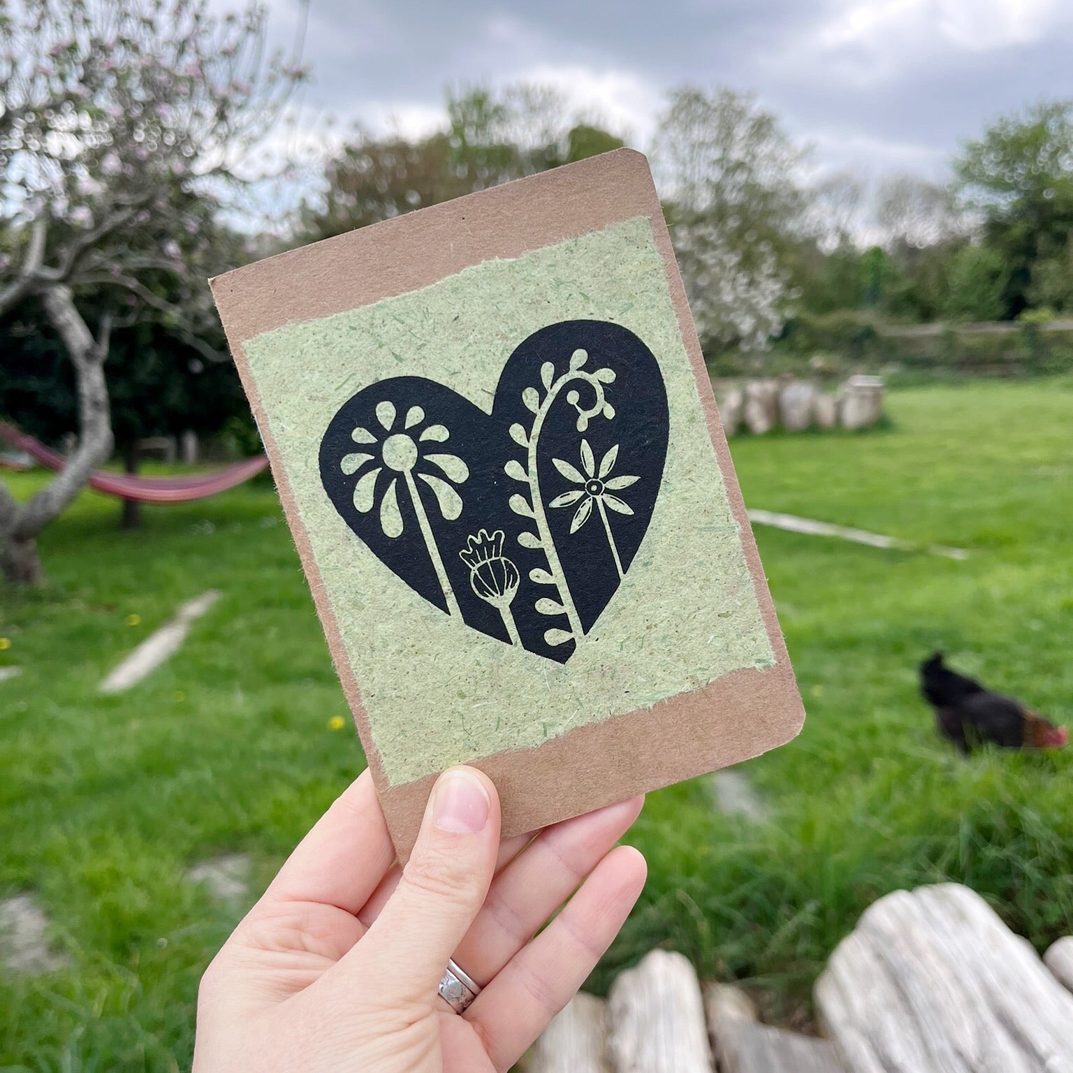 A6 kraft notebook with a black lino printed Floral Heart design on pale green handmade paper. Held in a hand in the garden.