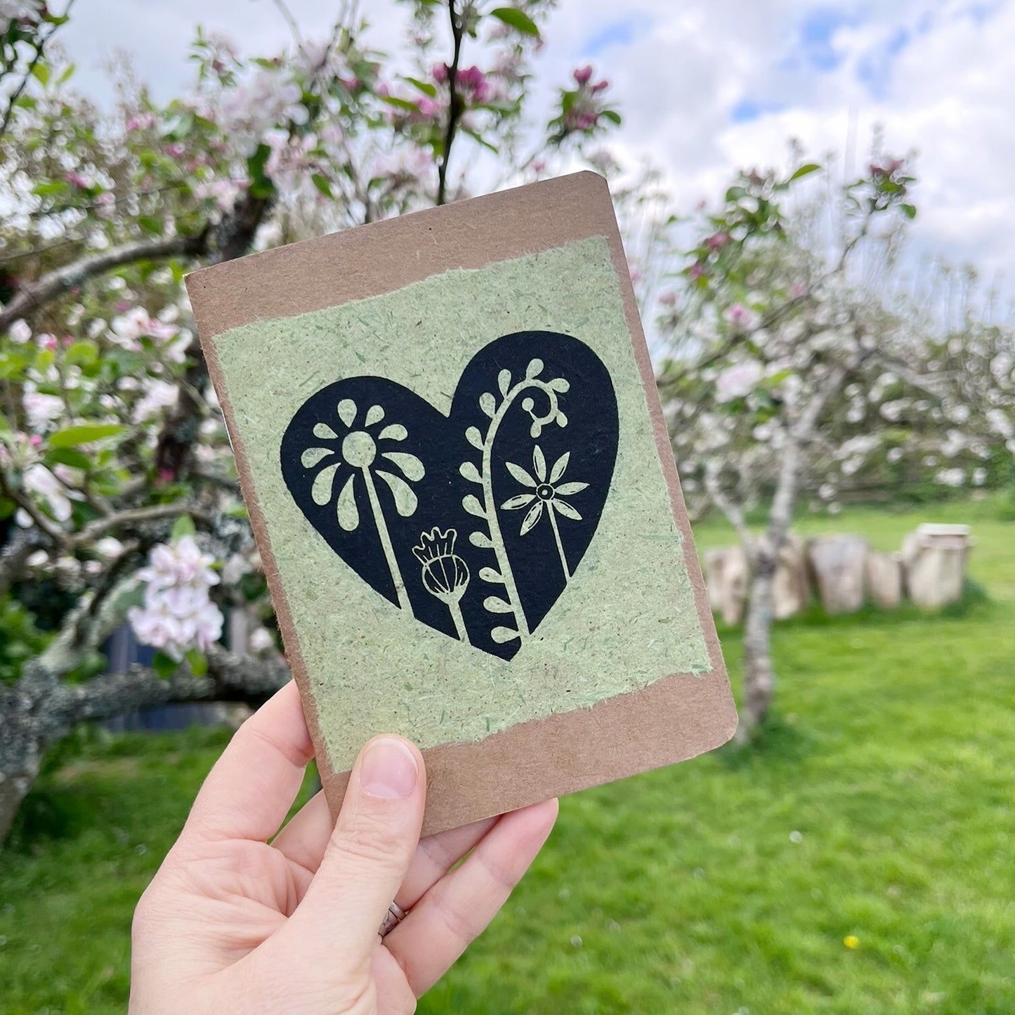 A6 kraft notebook with a black lino printed Floral Heart design on pale green handmade paper. Held in a hand in a garden.