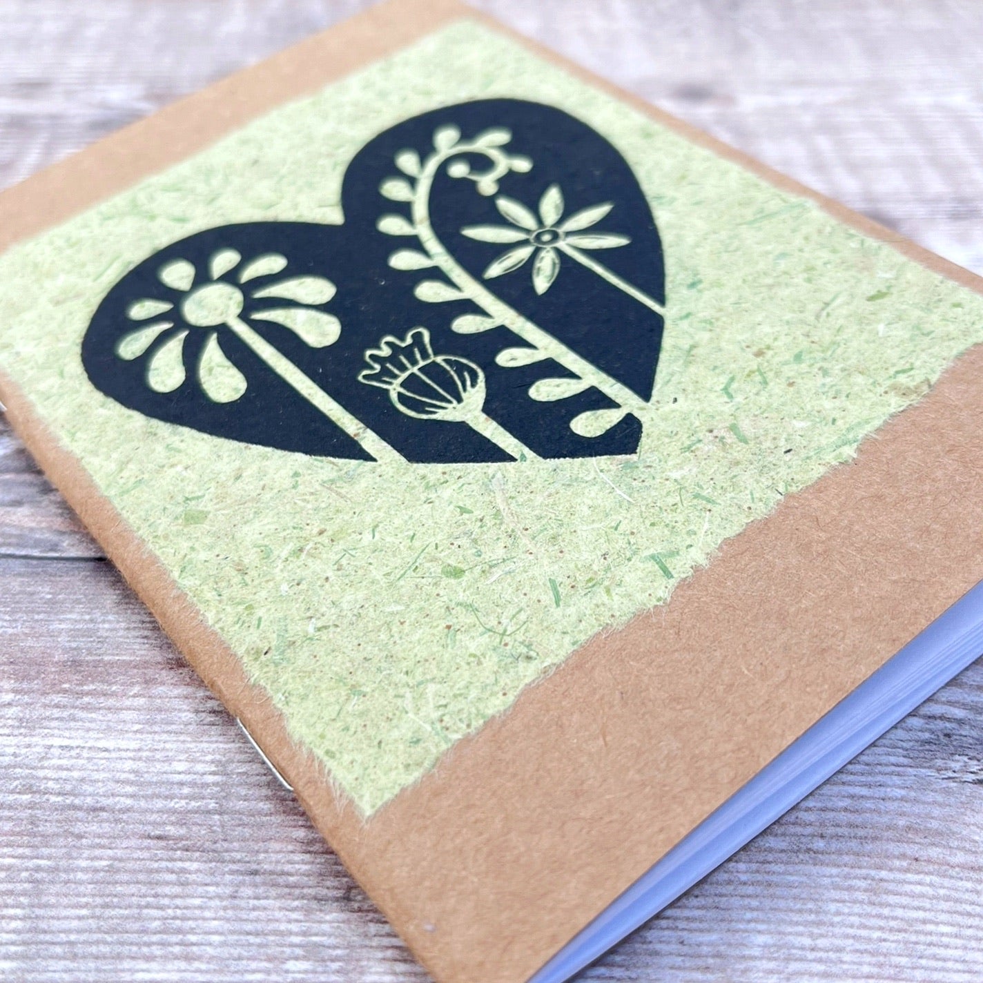 Close up of A6 kraft notebook with a black lino printed Floral Heart design on pale green handmade paper. Laid on a wooden background.