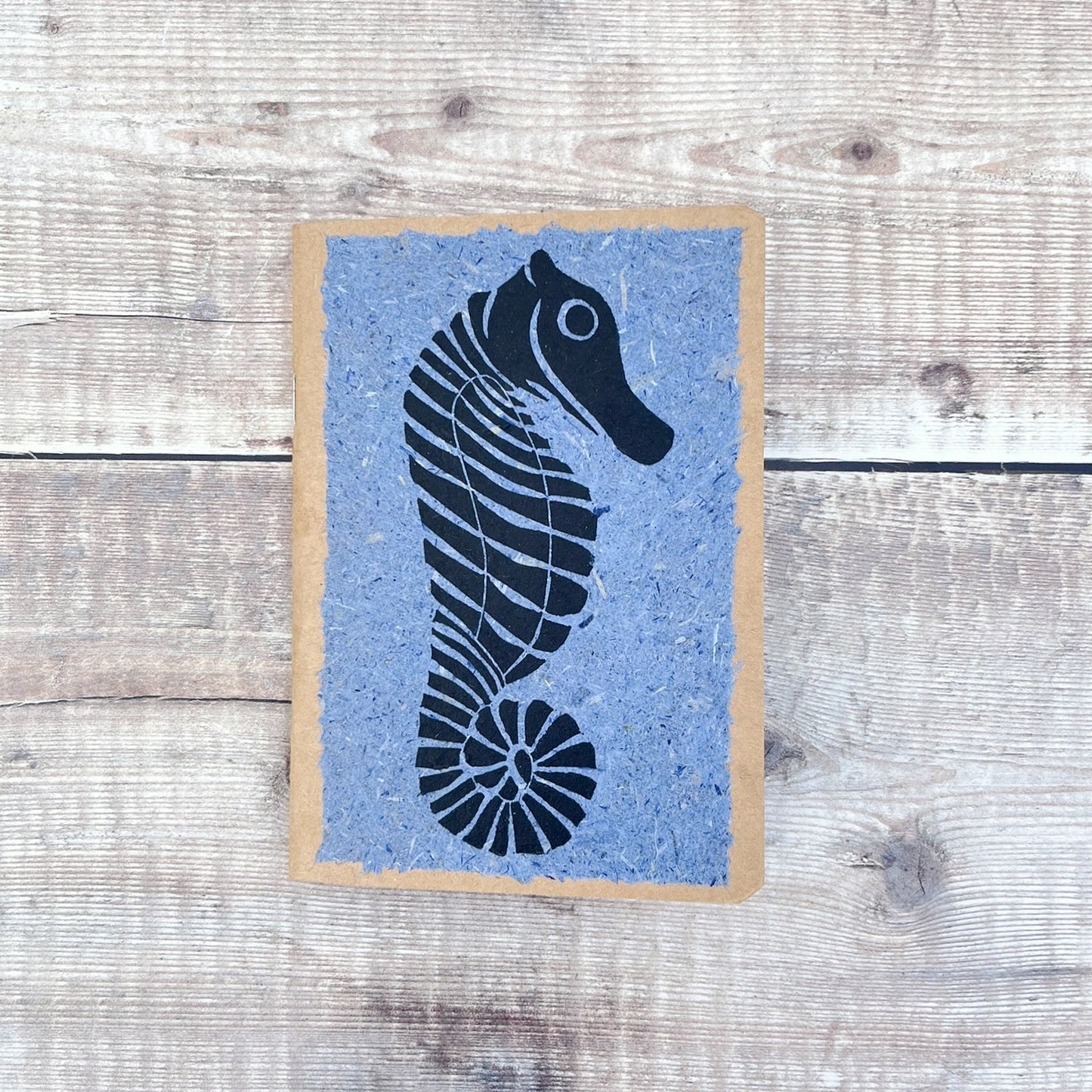 A6 kraft notebook with a black lino printed Seahorse on blue handmade paper. Laid on a wooden background.