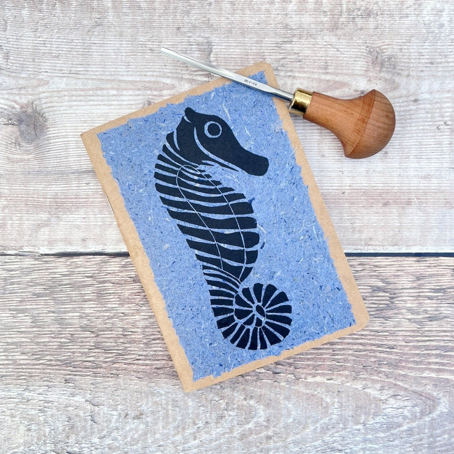 A6 kraft notebook with a black lino printed Seahorse on blue handmade paper. Photo includes the lino cutting tool used, laid on a wooden background.