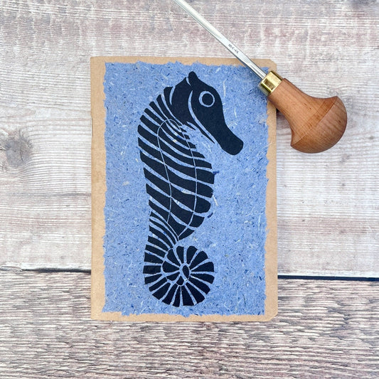 A6 kraft notebook with a black lino printed Seahorse on blue handmade paper. Photo includes the lino cutting tool used, laid on a wooden background.