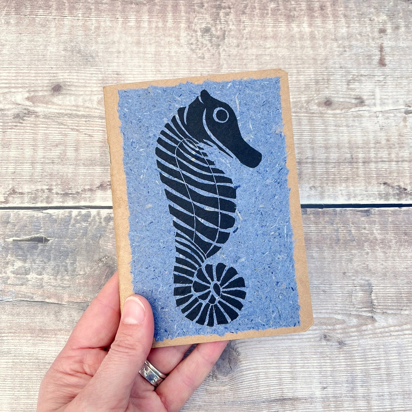 A6 kraft notebook with a black lino printed Seahorse on blue handmade paper. Held in a hand against a wooden background.