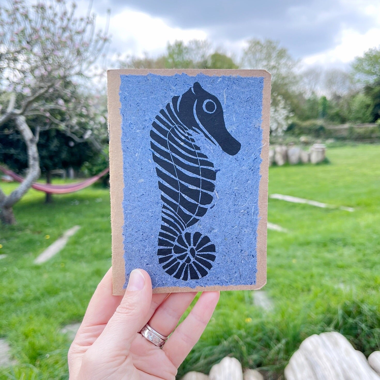 A6 kraft notebook with a black lino printed Seahorse on blue handmade paper. Held in a hand in a garden.