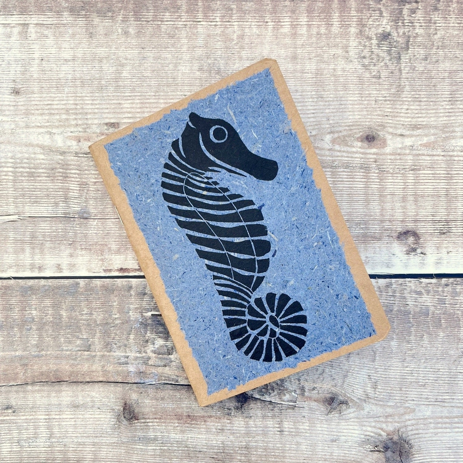A6 kraft notebook with a black lino printed Seahorse on blue handmade paper. Laid on a wooden background.
