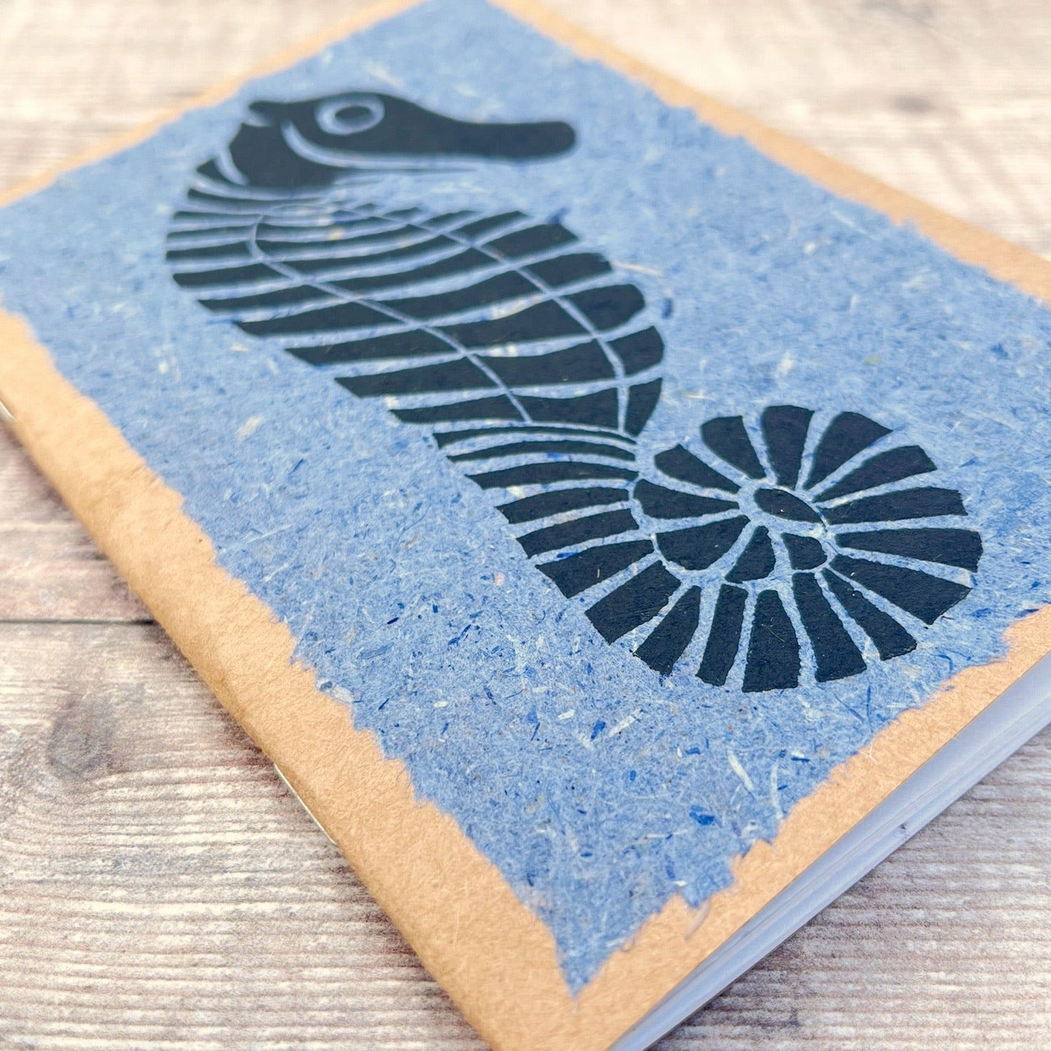 Close up of A6 kraft notebook with a black lino printed Seahorse on blue handmade paper. Laid on a wooden background.