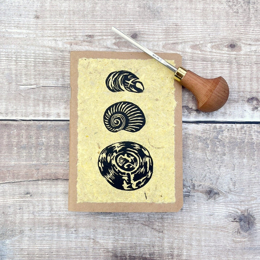 A6 kraft notebook with an original shell lino print in black ink on yellow handmade paper. Photo includes the lino cutting tool laid on a wooden background.