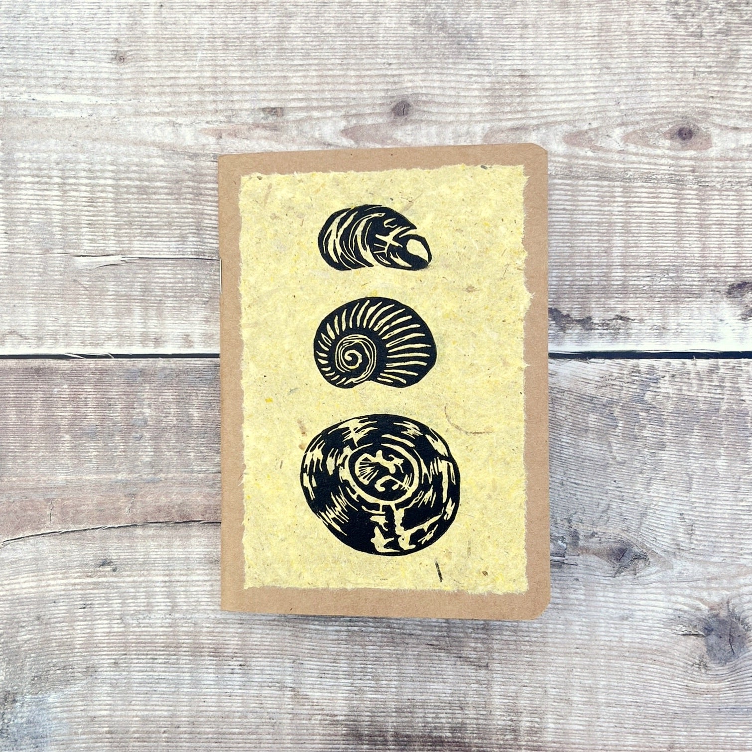A6 kraft notebook with an original shell lino print in black ink on yellow handmade paper. Laid on a wooden background.