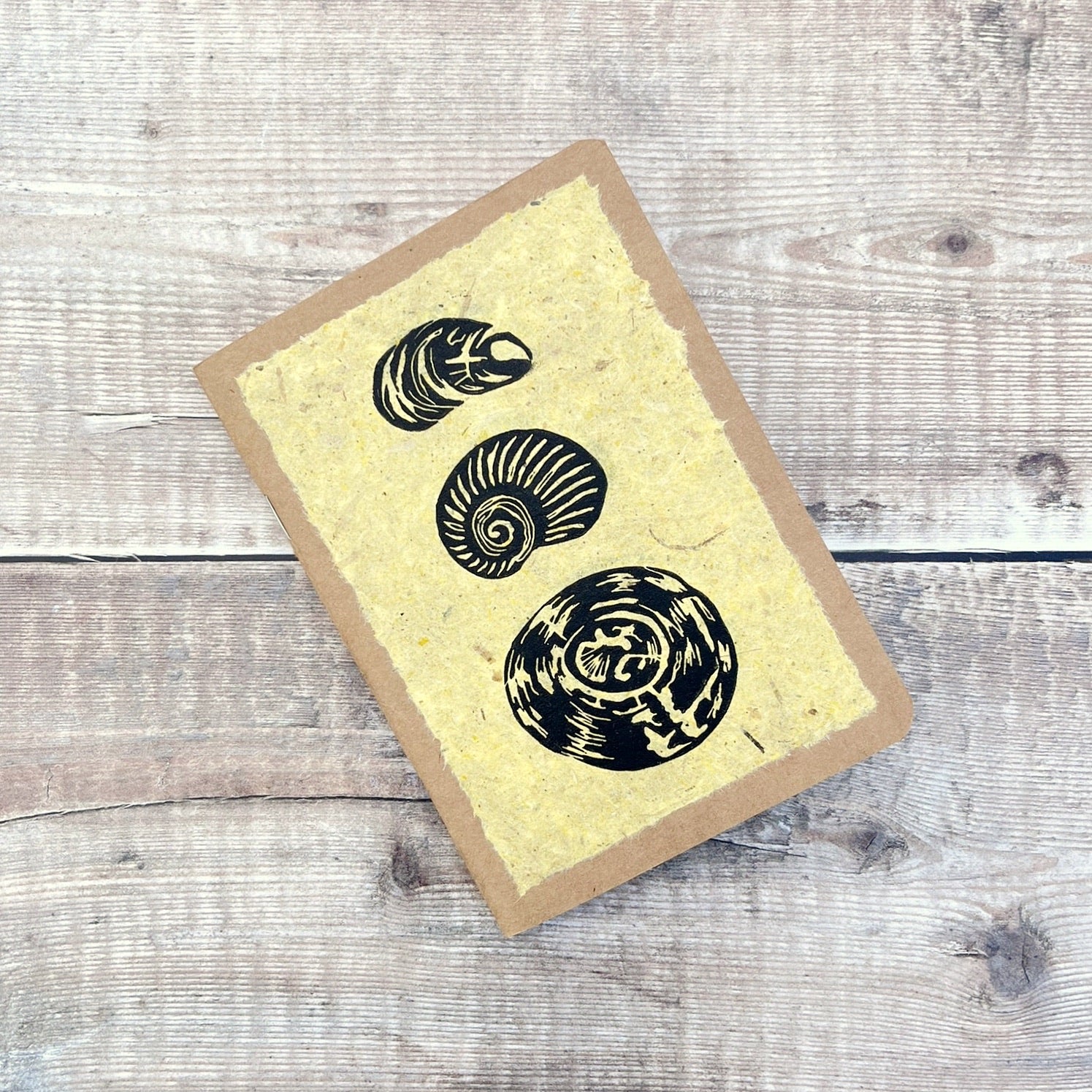 A6 kraft notebook with an original shell lino print in black ink on yellow handmade paper. Laid on a wooden background.