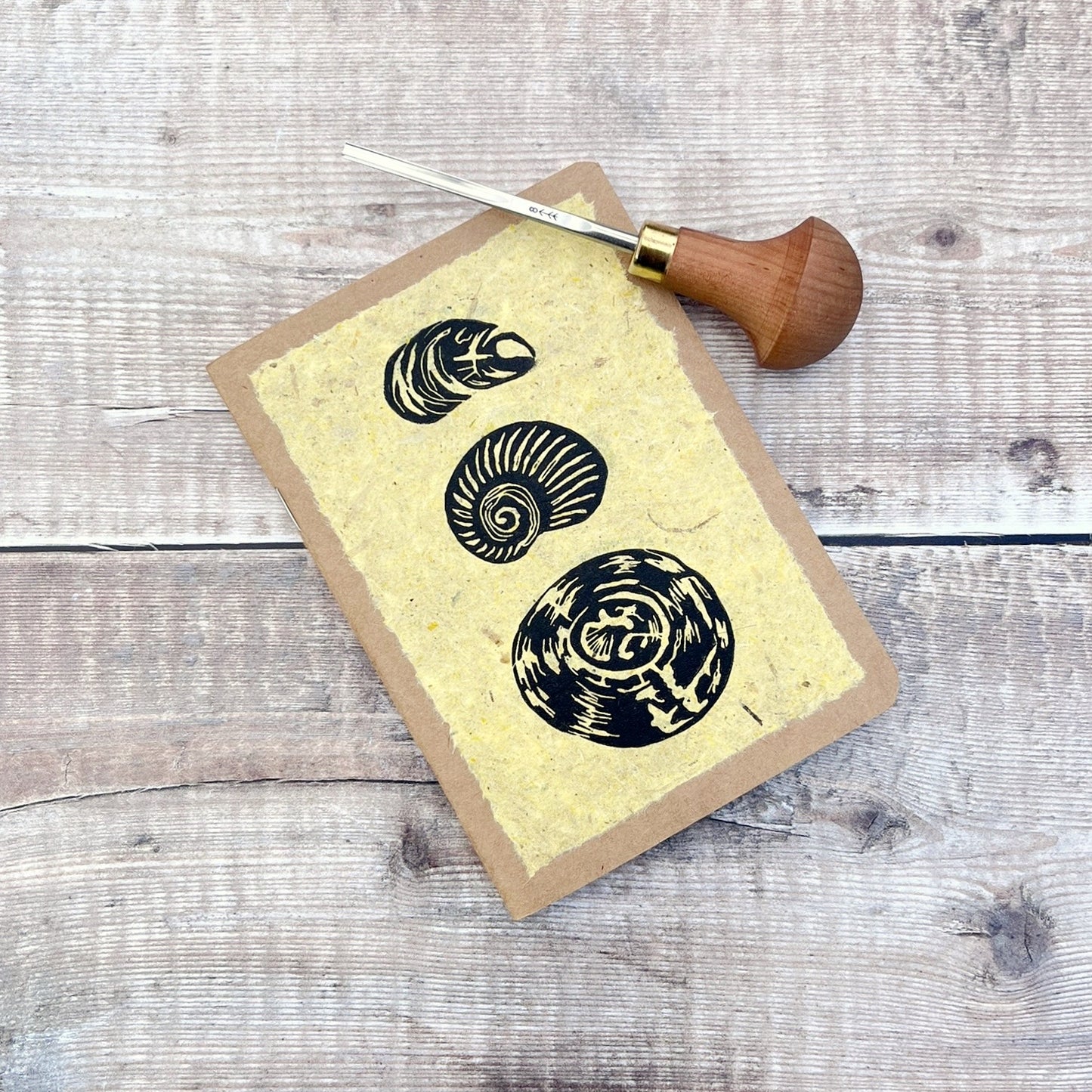 A6 kraft notebook with an original shell lino print in black ink on yellow handmade paper. Photo includes the lino cutting tool laid on a wooden background.