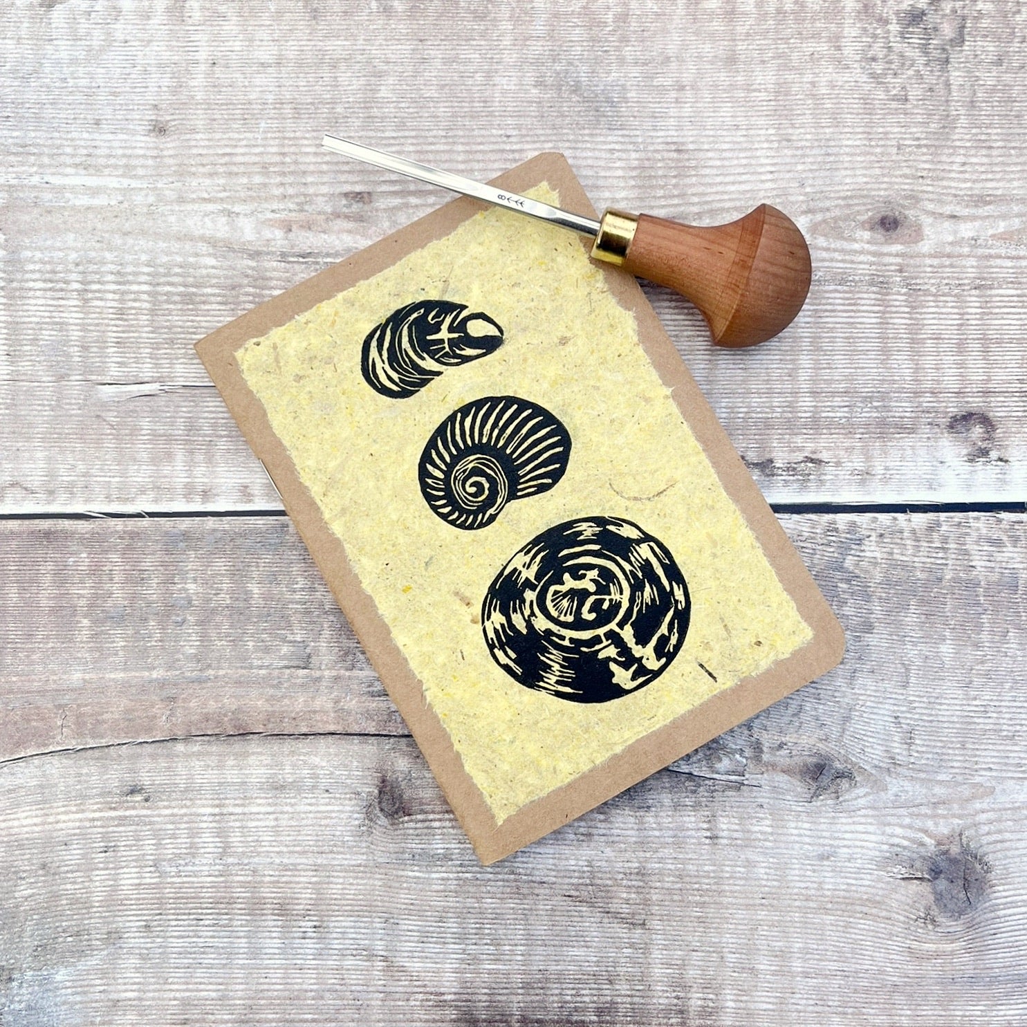 A6 kraft notebook with an original shell lino print in black ink on yellow handmade paper. Photo includes the lino cutting tool laid on a wooden background.