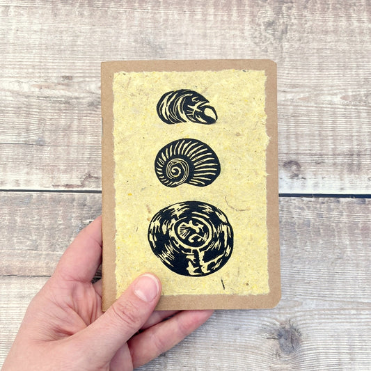 A6 kraft notebook with an original shell lino print in black ink on yellow handmade paper. Held in a hand against a wooden background.