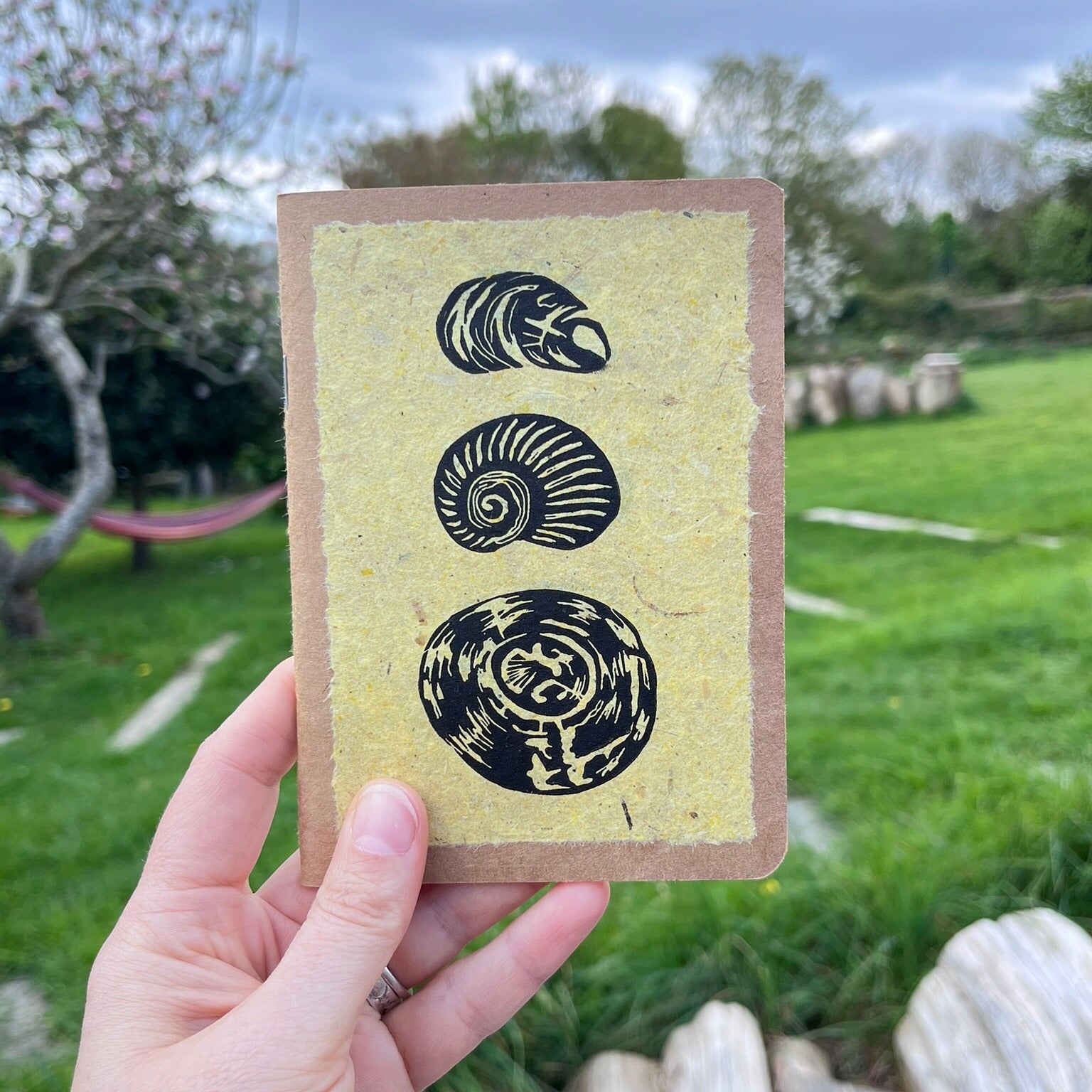 A6 kraft notebook with an original shell lino print in black ink on yellow handmade paper. Held in a hand in the garden.