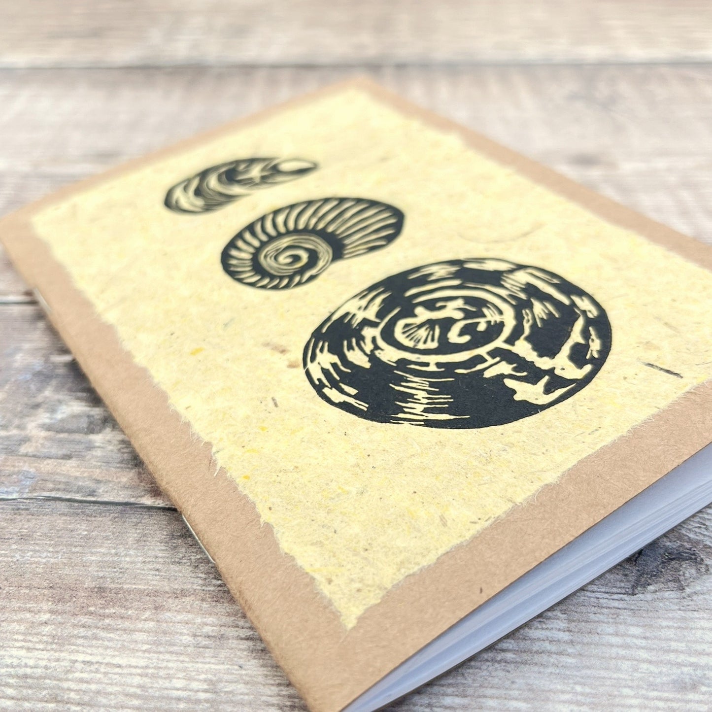 Close up of A6 kraft notebook with an original shell lino print in black ink on yellow handmade paper. Laid on a wooden background.