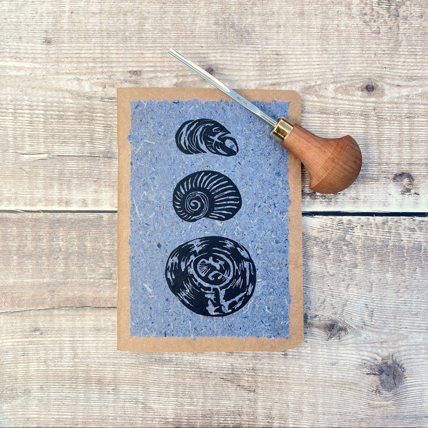 A6 kraft notebook with black lino printed Seashells on blue handmade paper. Photo includes the lino cutting tool used, laid on a wooden background.
