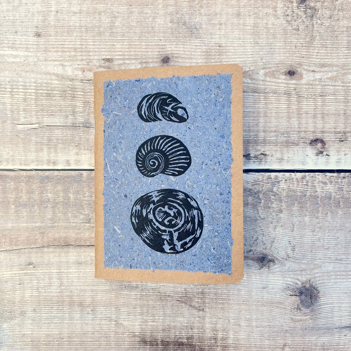 A6 kraft notebook with black lino printed Seashells on blue handmade paper. Laid on a wooden background.