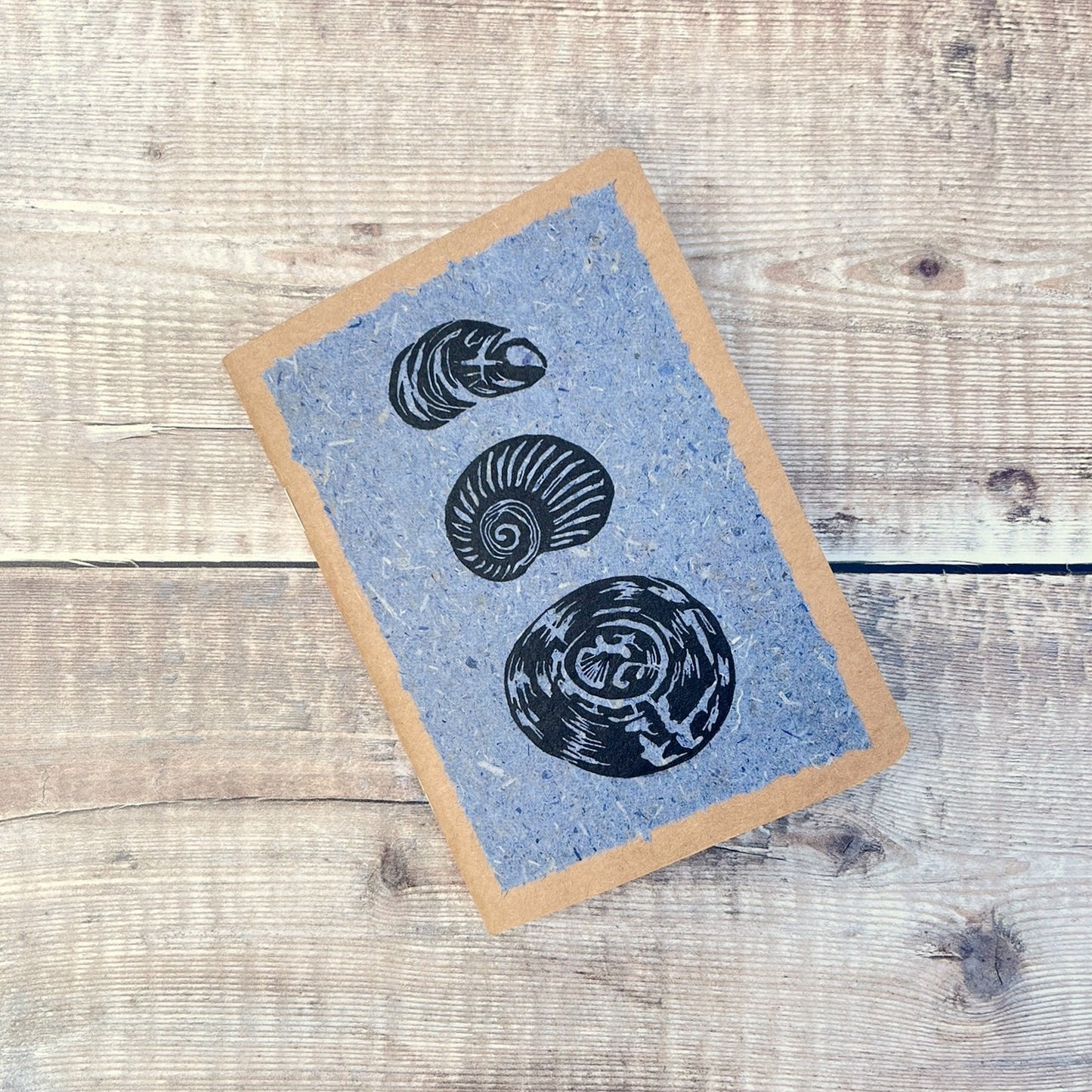 A6 kraft notebook with black lino printed Seashells on blue handmade paper. Laid on a wooden background.