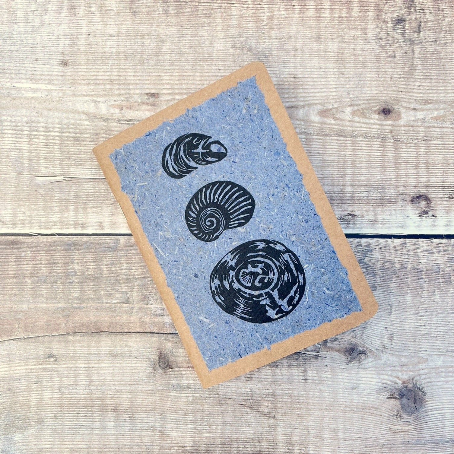 A6 kraft notebook with black lino printed Seashells on blue handmade paper. Laid on a wooden background.