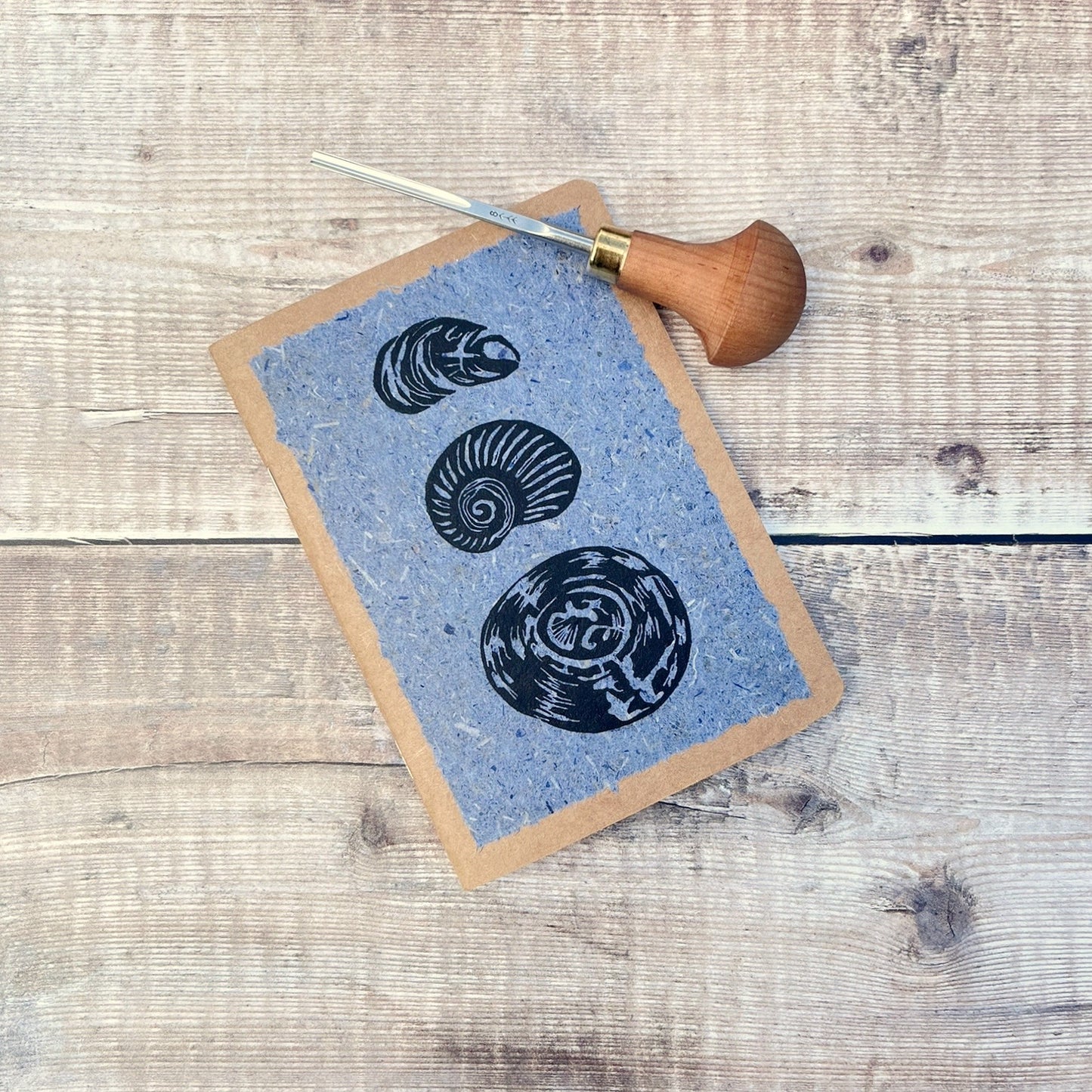 A6 kraft notebook with black lino printed Seashells on blue handmade paper. Photo includes the lino cutting tool used, laid on a wooden background.