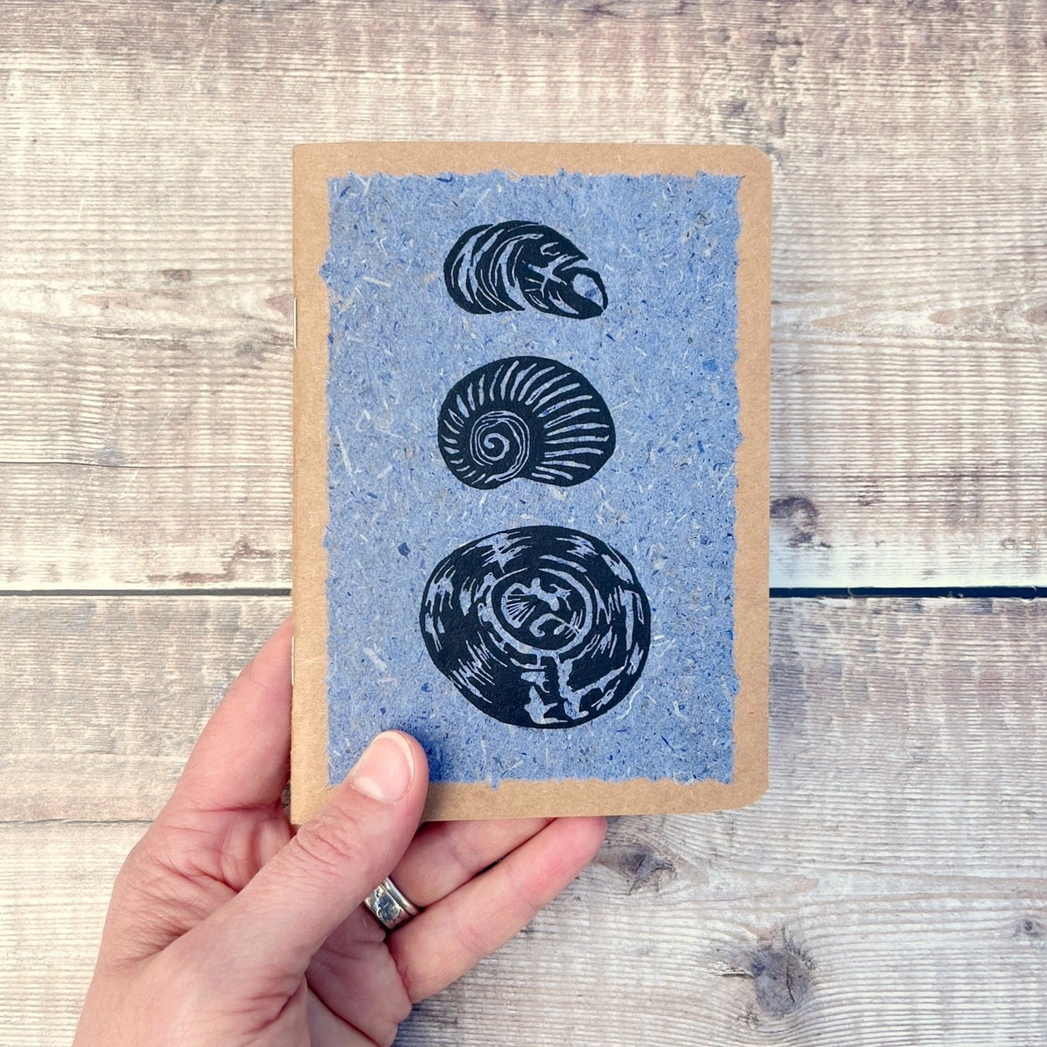 A6 kraft notebook with black lino printed Seashells on blue handmade paper. Held in a hand against a wooden background.