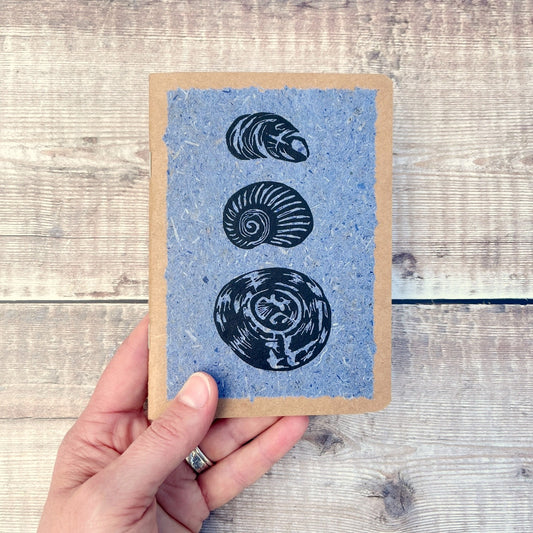 A6 kraft notebook with black lino printed Seashells on blue handmade paper. Held in a hand against a wooden background.