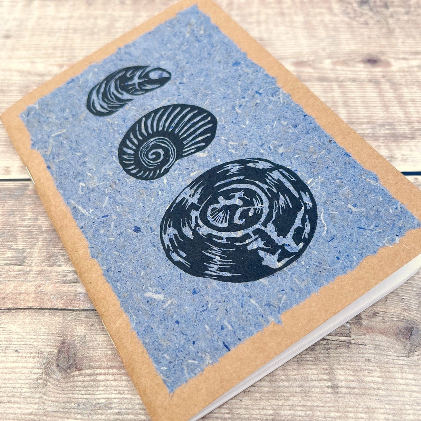 Close up of A6 kraft notebook with black lino printed Seashells on blue handmade paper. Laid on a wooden background.
