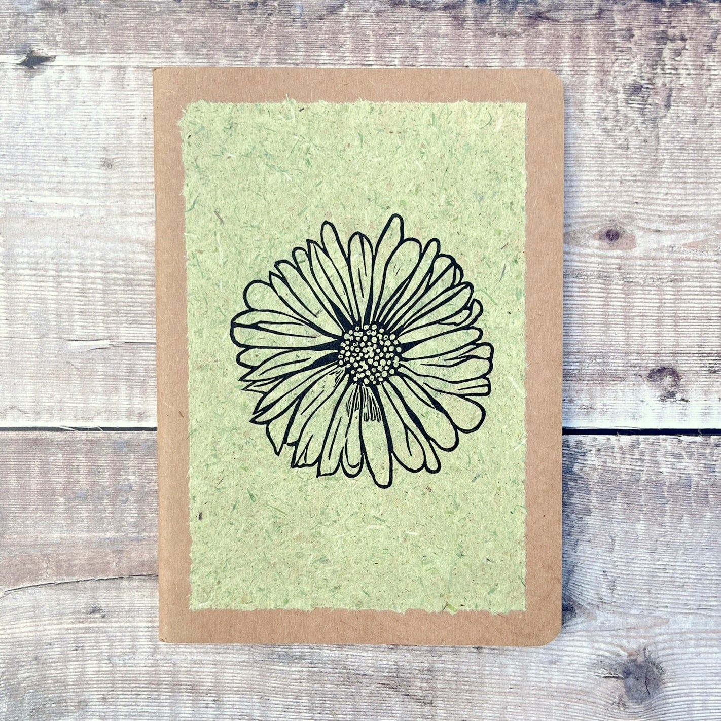 A5 kraft sketchbook with an original lino print of a Daisy on pale green handmade paper. Laid on a wooden background.