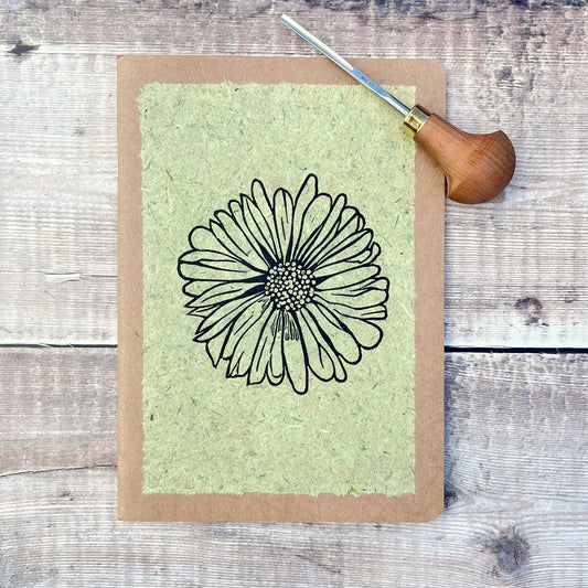 A5 kraft sketchbook with an original lino print of a Daisy on pale green handmade paper. Photo includes lino cutting tool laid on a wooden background.