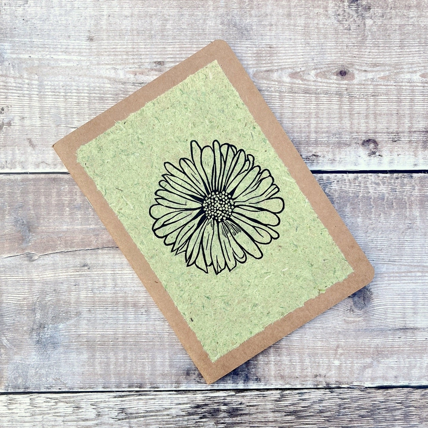 A5 kraft sketchbook with an original lino print of a Daisy on pale green handmade paper. Laid on a wooden background.