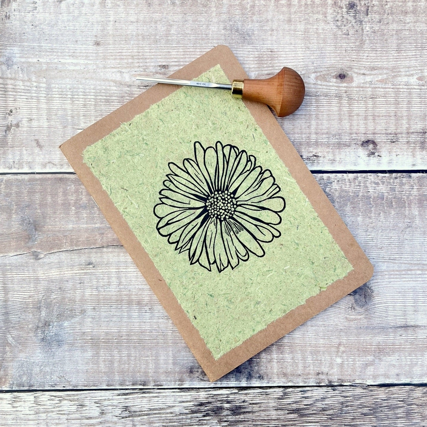 A5 kraft sketchbook with an original lino print of a Daisy on pale green handmade paper. Photo includes lino cutting tool laid on a wooden background.