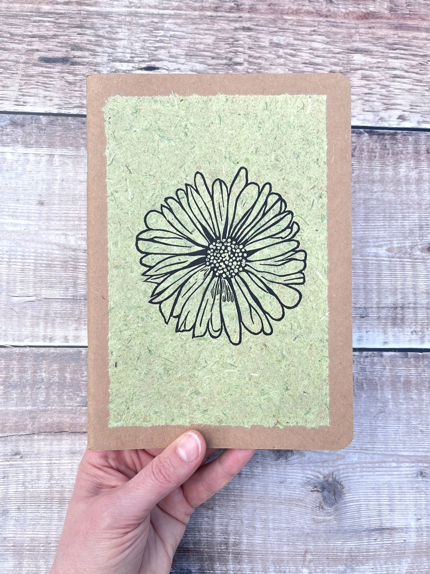 A5 kraft sketchbook with an original lino print of a Daisy on pale green handmade paper. Held in a hand against a wooden background.