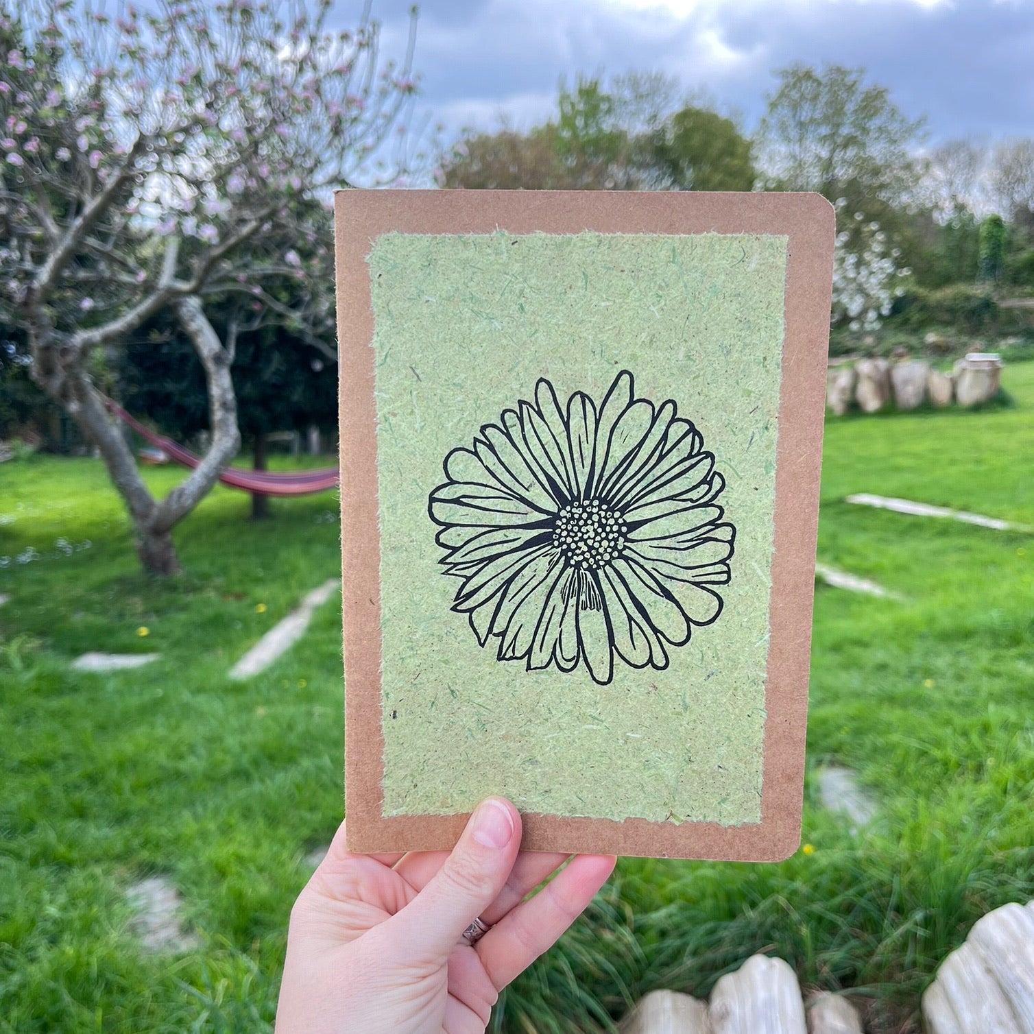 A5 kraft sketchbook with an original lino print of a Daisy on pale green handmade paper. Held in a hand in a garden.