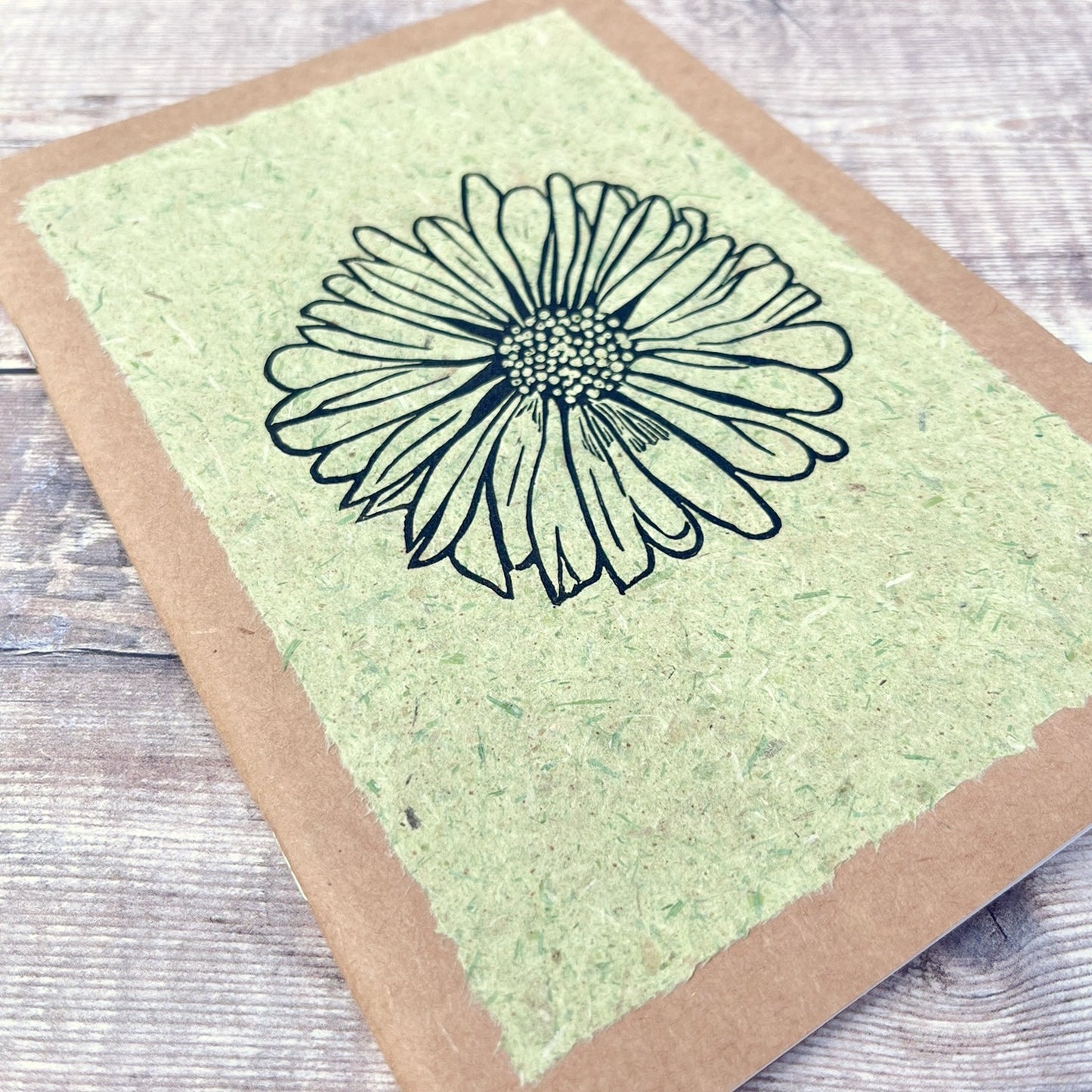 Close up of A5 kraft sketchbook with an original lino print of a Daisy on pale green handmade paper. Laid on a wooden background.