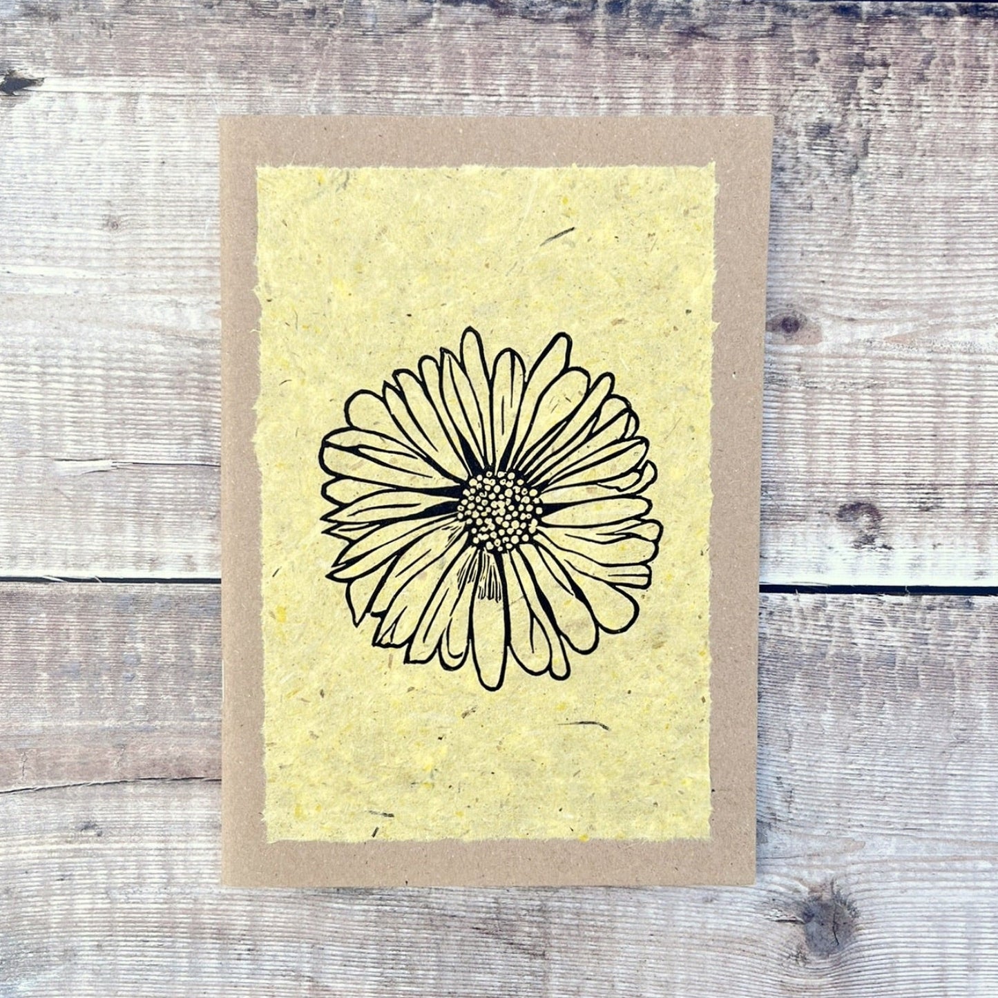 A5 kraft sketchbook with an original lino print of a Daisy on handmade yellow paper. Laid on a wooden background.