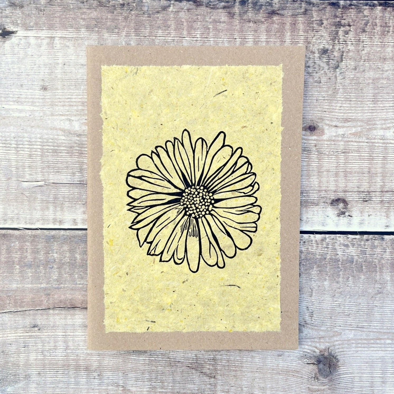A5 kraft sketchbook with an original lino print of a Daisy on handmade yellow paper. Laid on a wooden background.