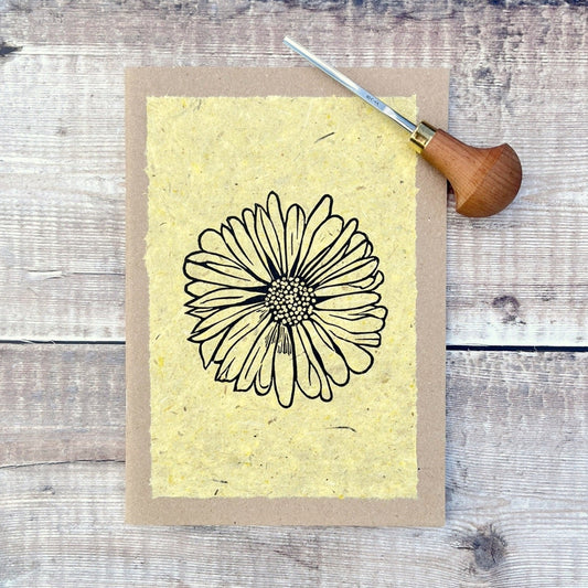 A5 kraft sketchbook with an original lino print of a Daisy on handmade yellow paper. Photo includes lino cutting tool laid on a wooden background.