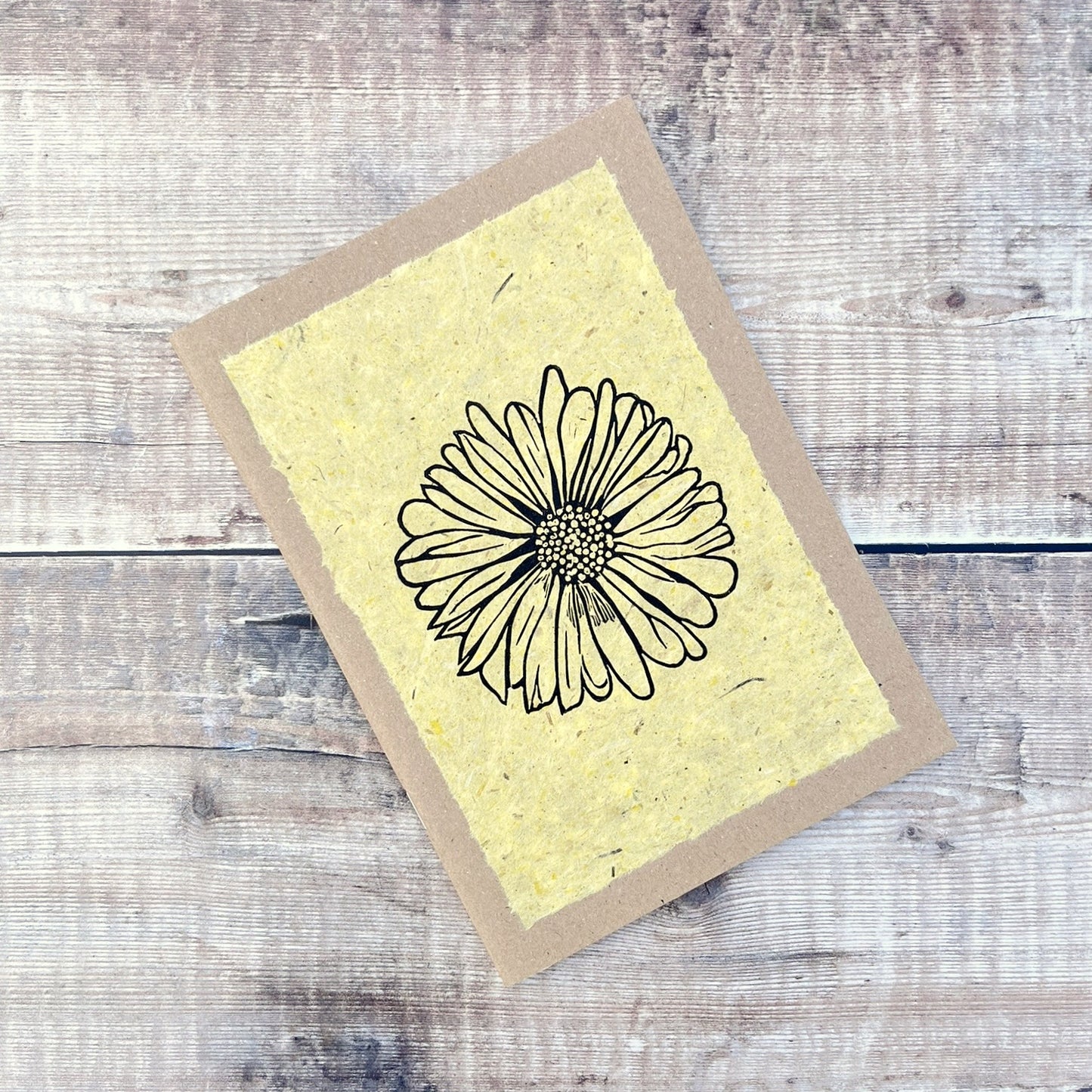 A5 kraft sketchbook with an original lino print of a Daisy on handmade yellow paper. Laid on a wooden background.