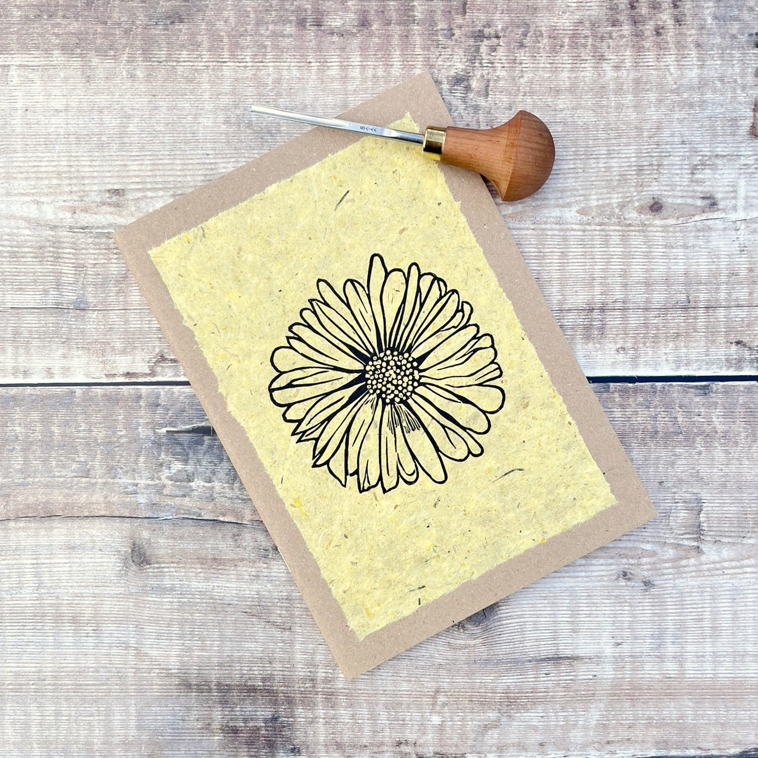 A5 kraft sketchbook with an original lino print of a Daisy on handmade yellow paper. Photo includes lino cutting tool laid on a wooden background.