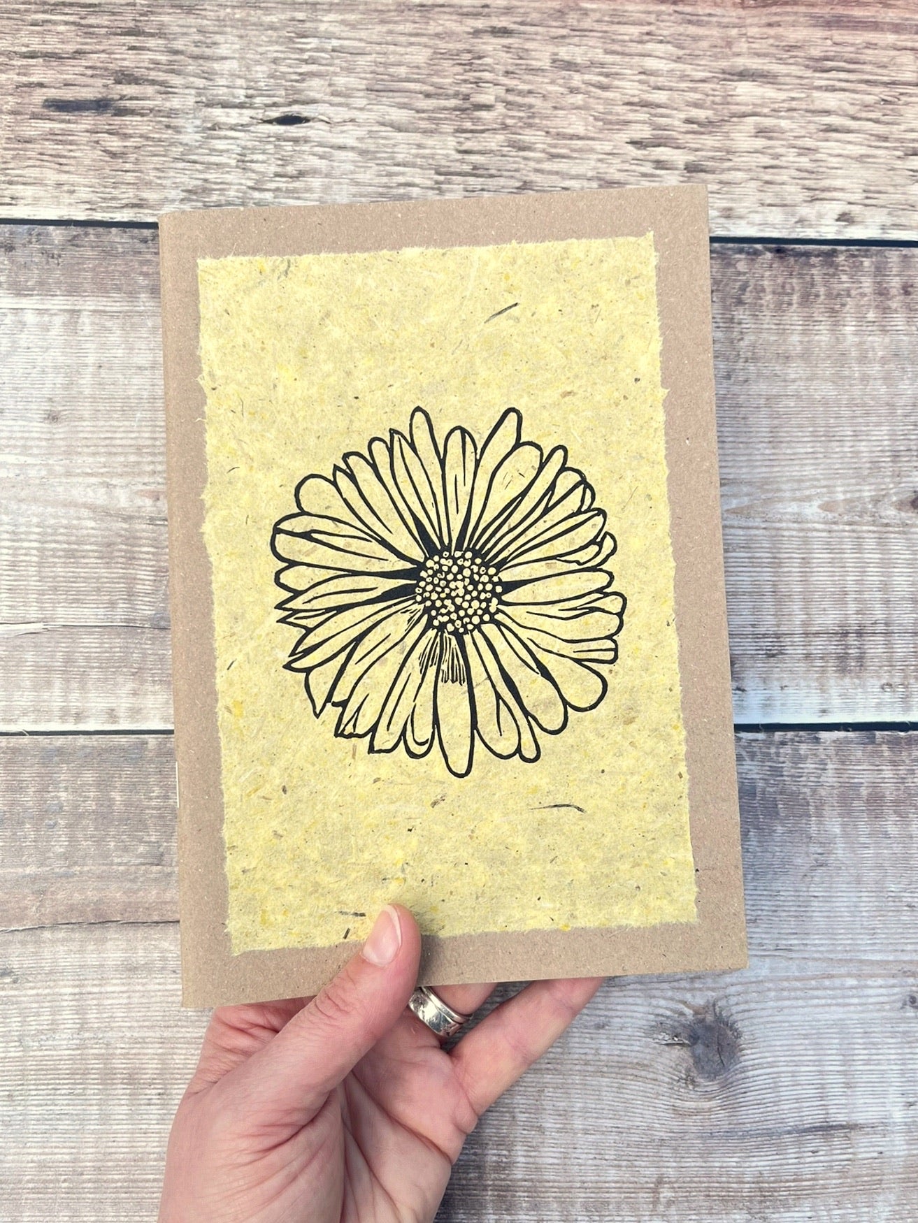 A5 kraft sketchbook with an original lino print of a Daisy on handmade yellow paper. Held in a hand against a wooden background.