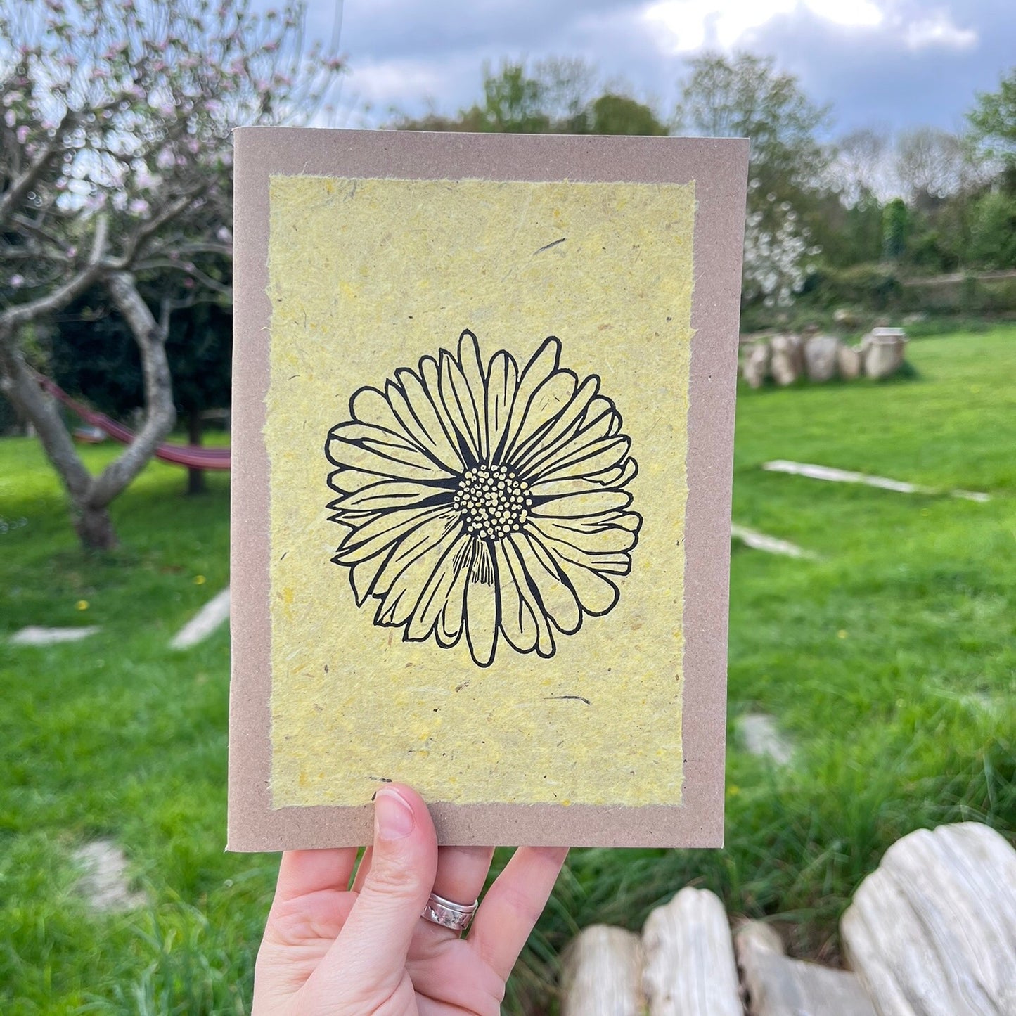A5 kraft sketchbook with an original lino print of a Daisy on handmade yellow paper. Held in a hand in the garden.