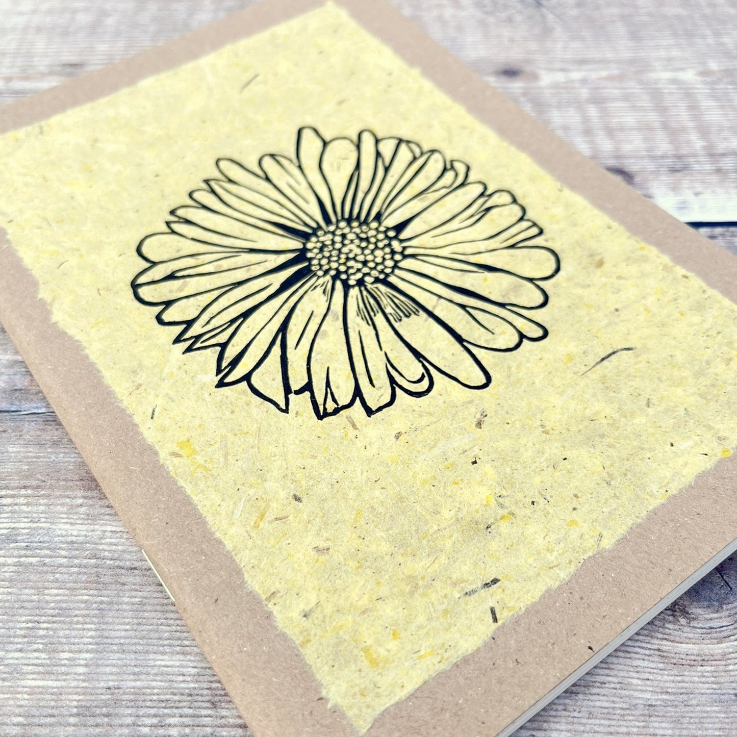 Close up of A5 kraft sketchbook with an original lino print of a Daisy on handmade yellow paper. Laid on a wooden background.