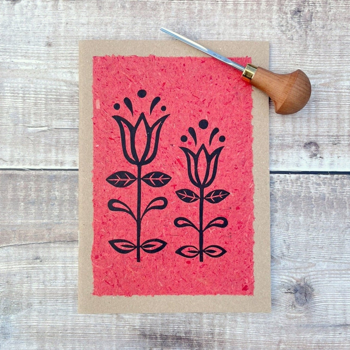 A5 kraft sketchbook with an original lino print of Tulips on handmade brick red paper. Photo includes lino cutting tool laid on a wooden background.