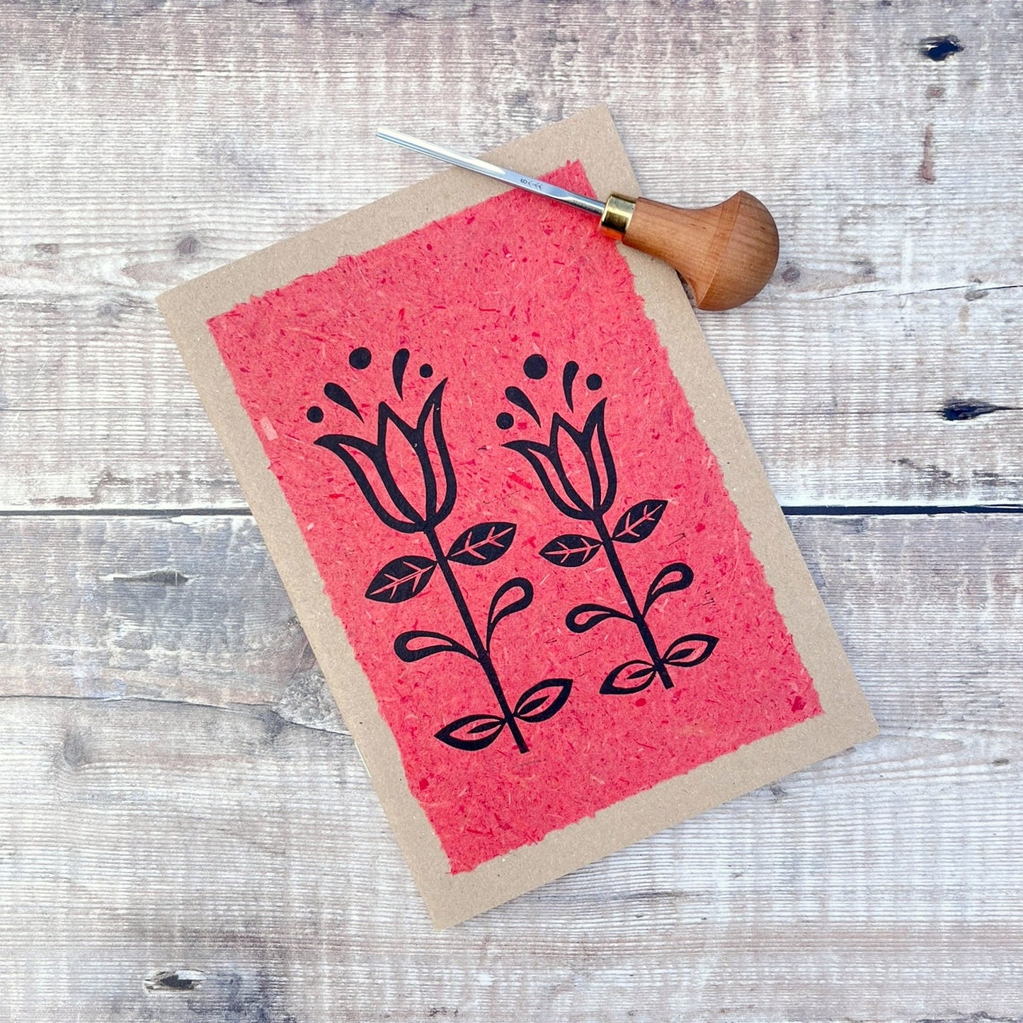 A5 kraft sketchbook with an original lino print of Tulips on handmade brick red paper. Photo includes lino cutting tool laid on a wooden background.